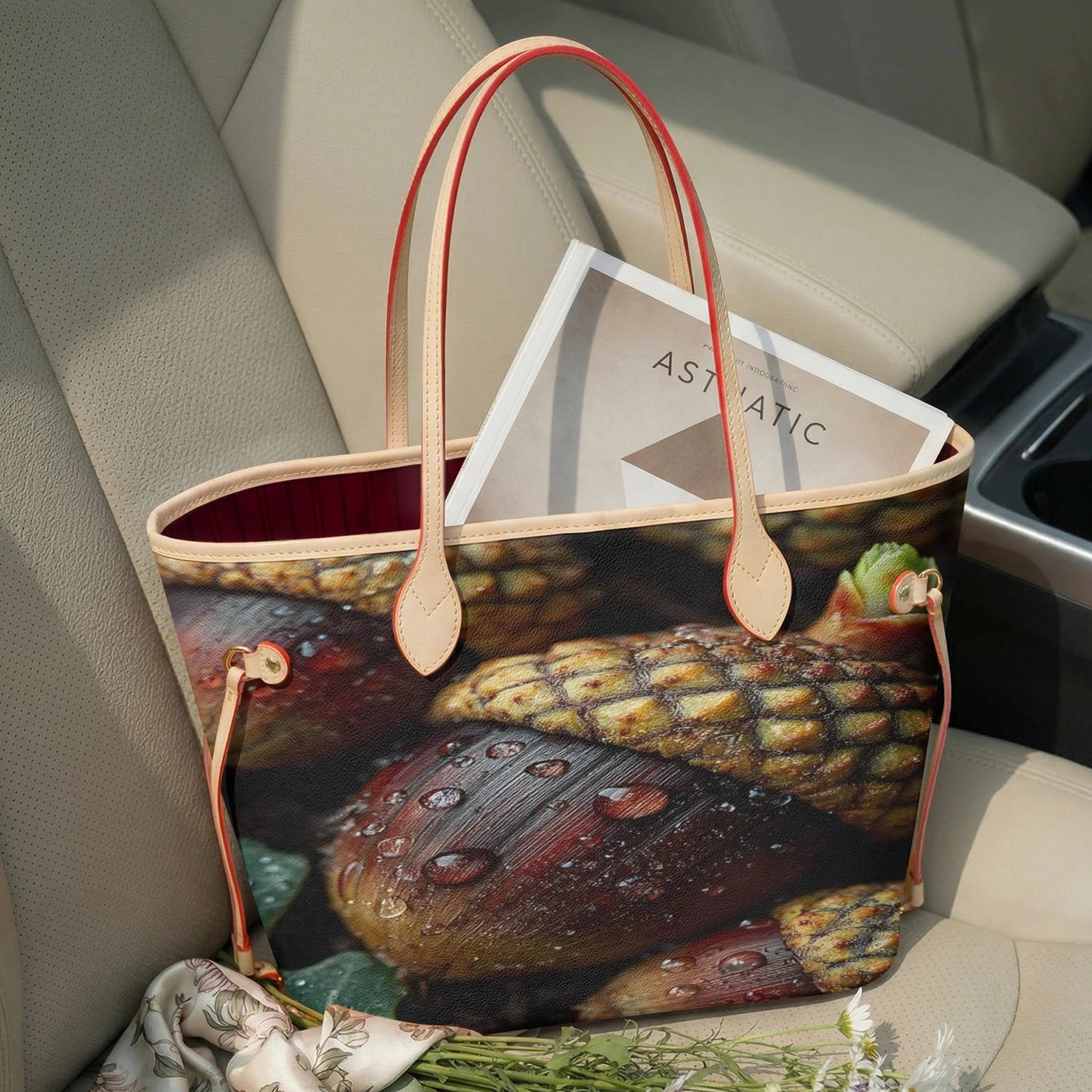 A tote bag with a realistic acorn print sits on a beige car seat. The bag has light tan leather handles and trim, and a red interior lining.