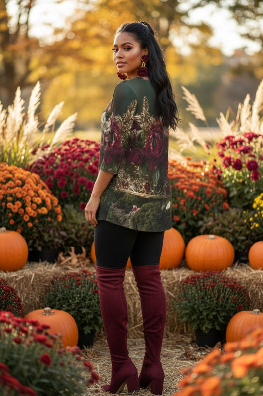 A woman stands in an autumn garden setting surrounded by pumpkins and colorful flowers, wearing a dark green and burgundy floral print tunic top, black leggings, and burgundy over-the-knee boots.