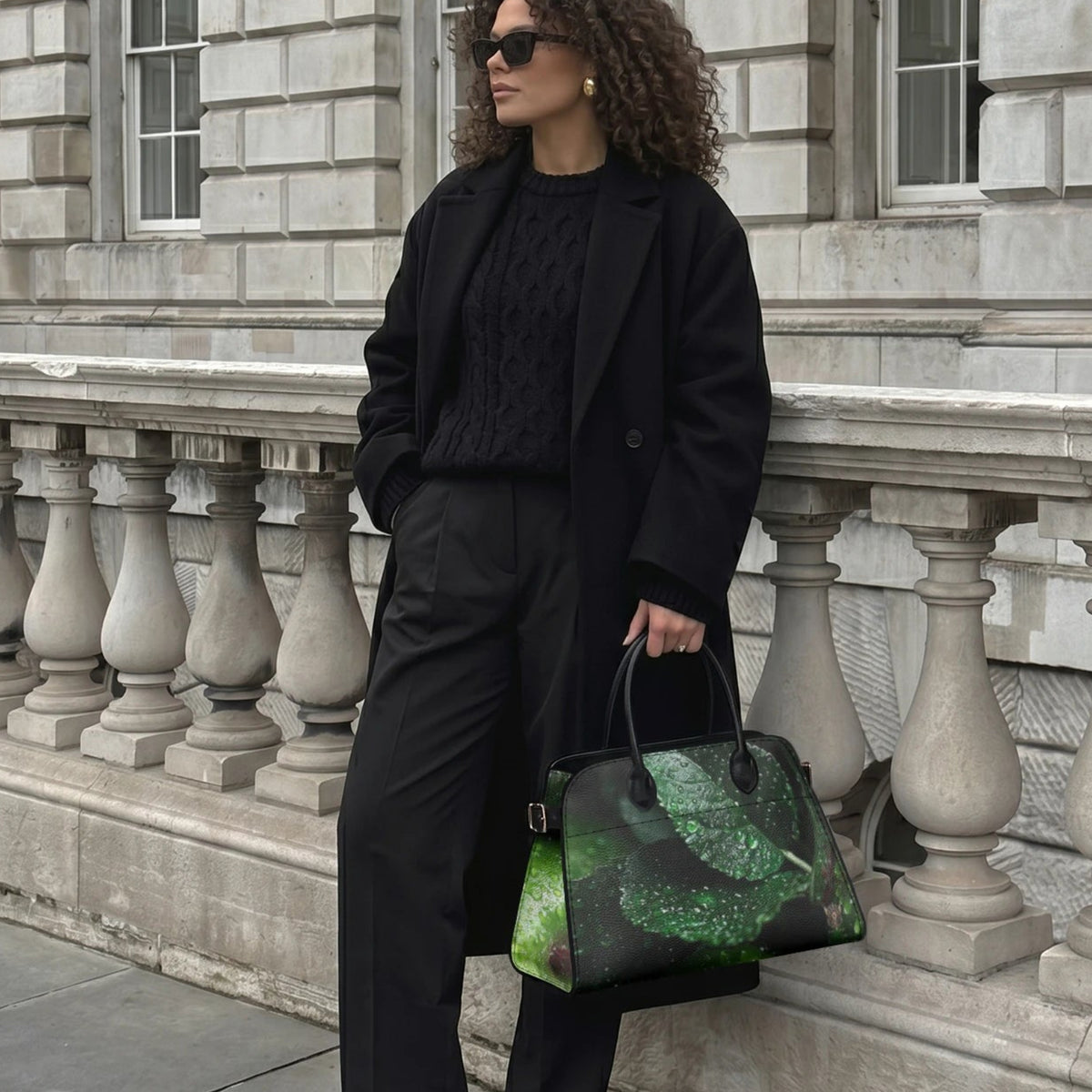 A woman stands on a stone sidewalk in front of a gray stone building, wearing a black coat, black top, and black trousers while holding a black tote bag with a green and gold leaf pattern.