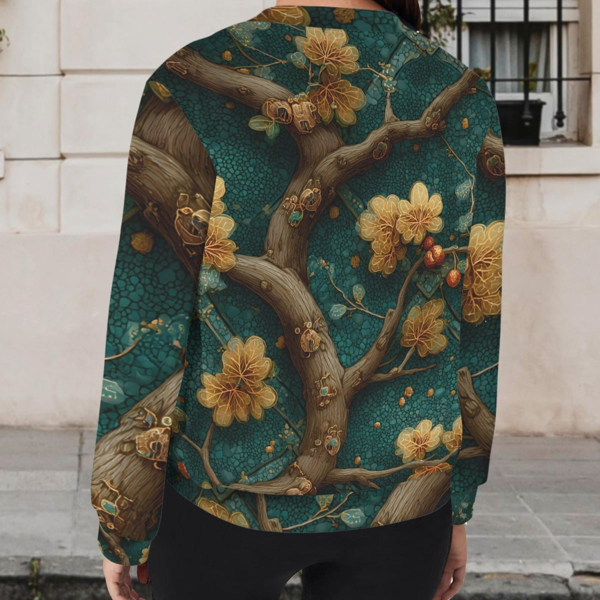 A woman viewed from behind wearing a long-sleeve bomber-style jacket with an intricate floral and branch print in teal, gold, and brown, standing outdoors in front of a light-colored building wall.