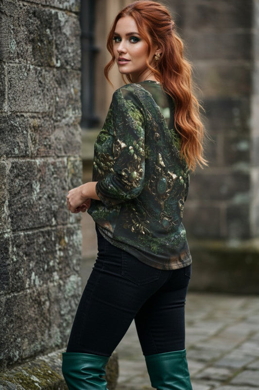 A woman with red hair stands in front of a stone wall wearing a dark green camouflage-patterned long-sleeve top, black pants, and dark green knee-high boots.