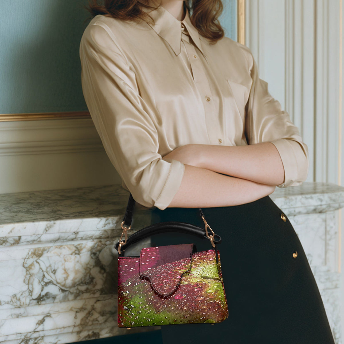 A woman wearing a beige satin shirt and black skirt stands in front of a marble mantel, carrying a small structured handbag with a glittery multicolor pattern in pink, green, and gold.