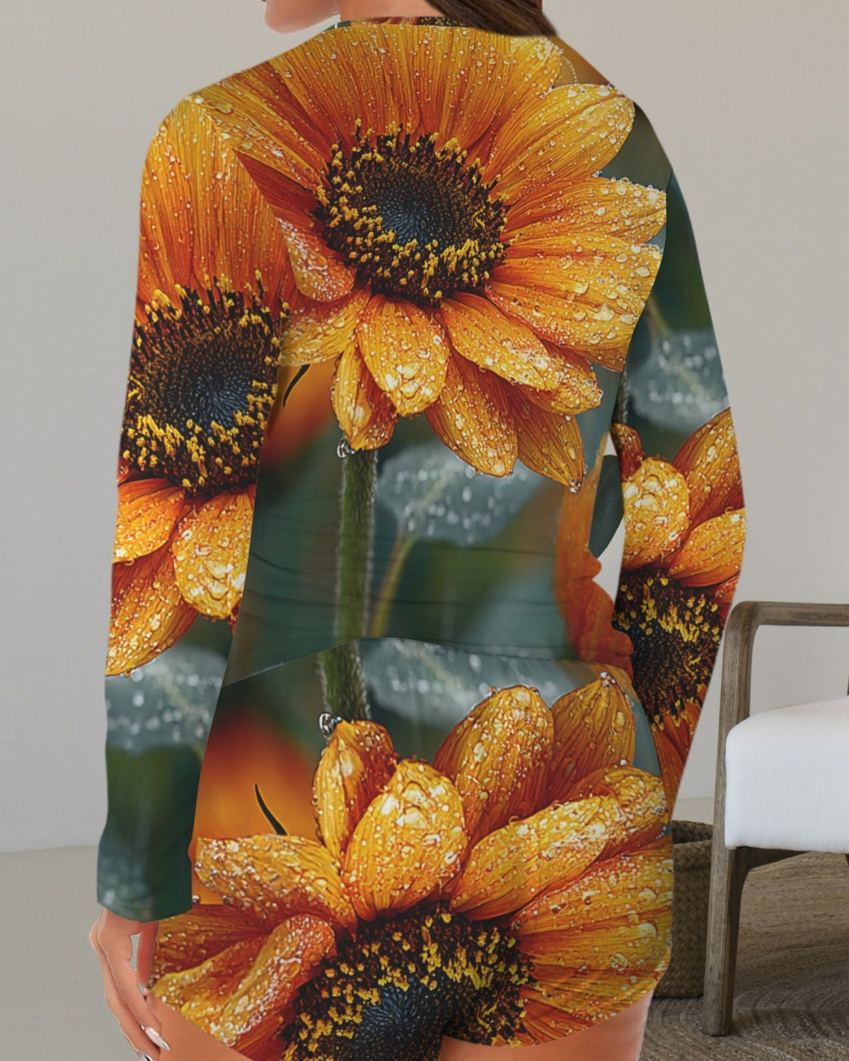 A woman wearing a long-sleeve romper with a vibrant sunflower print, shown from the back in an indoor setting with a white armchair.