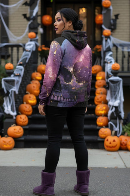 A woman stands in front of a Halloween-decorated house doorway, wearing a purple and black hooded sweatshirt with a large skull and galaxy print, black leggings, and purple boots.