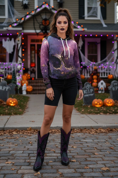 A woman stands on a cobblestone street in front of a Halloween-decorated house, wearing a purple and black galaxy-print hoodie with white drawstrings, black biker shorts, and dark purple cowboy boots.
