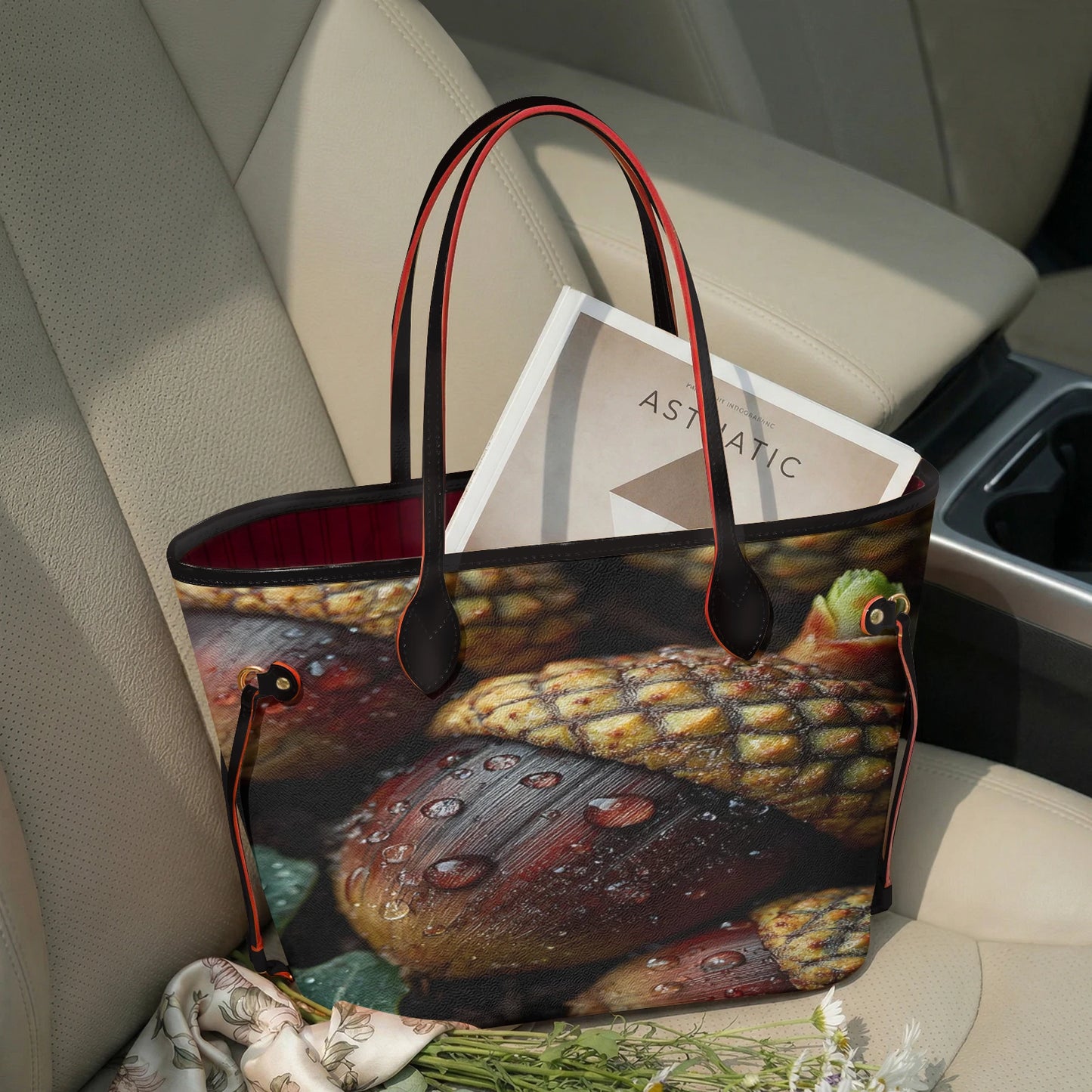 A tote bag with a realistic acorn print sits on a beige car seat. The bag features dark brown and black acorns with water droplets, a red interior lining, and red trim on the handles.