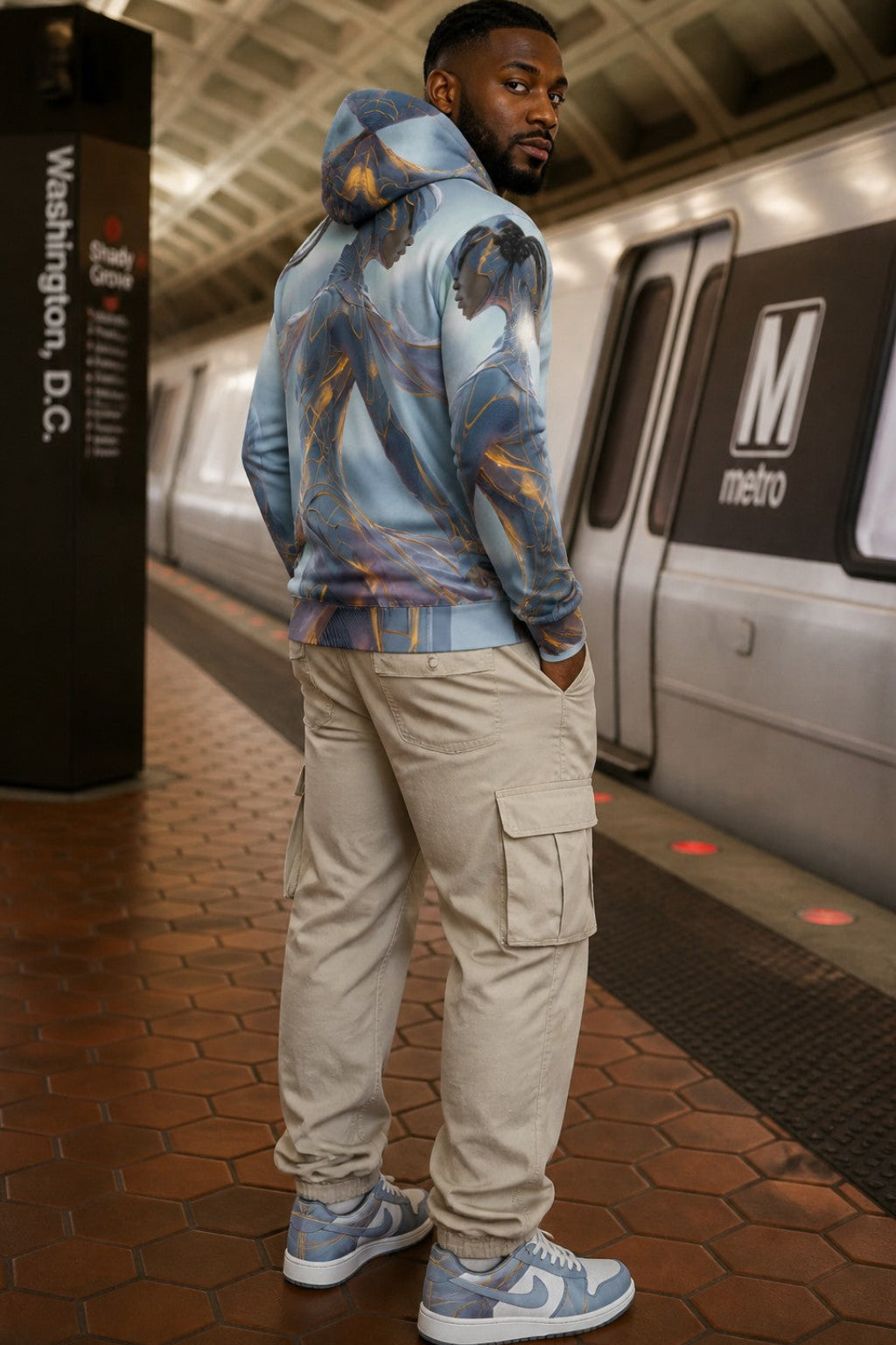 A man wearing a light blue and gray abstract-patterned hoodie, beige cargo pants, and light blue and gray sneakers stands on a Washington, D.C.