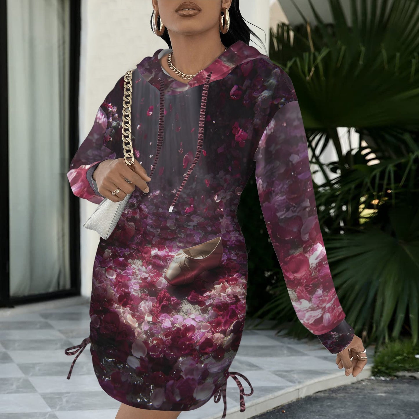 A woman wearing a long-sleeve hooded dress with a dark purple and pink floral print, holding a white handbag with a gold chain strap in an outdoor patio setting.