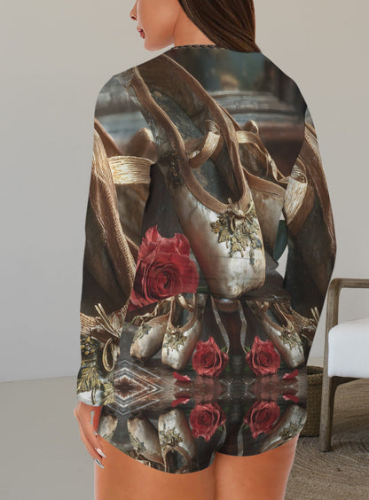 A woman shown from the back wearing a long-sleeve romper with an intricate vintage-style print featuring a large metallic bag, gold chains, and red roses. The background shows a modern white armchair in a minimalist room.