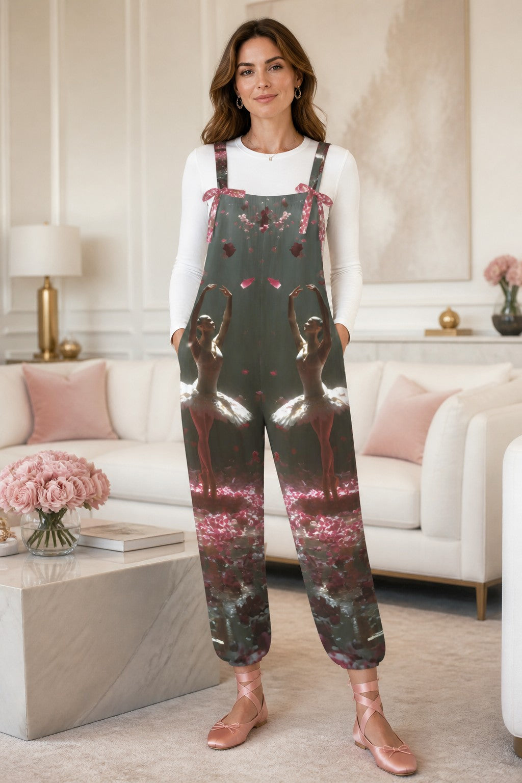 A woman modeling a pair of dark green overall-style pants with a pink flamingo and floral print, worn over a white long-sleeve top in a bright, modern living room.