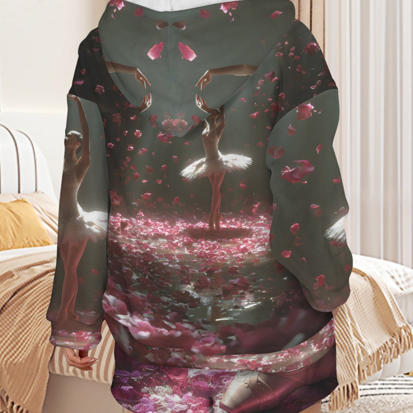 A person shown from the back wearing a long hooded sweatshirt with an all-over fantasy-style print featuring ballerinas, pink petals, and a dark green base, photographed in a bedroom setting.