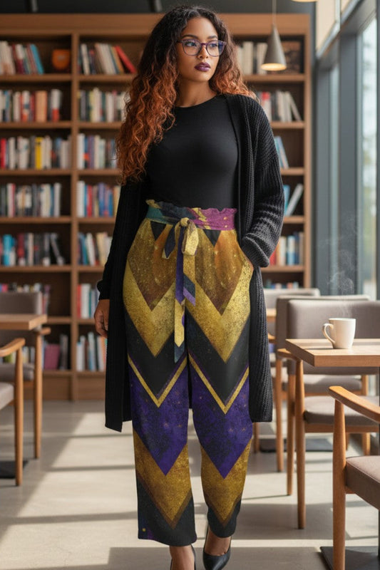 A woman stands in a modern library or cafe setting, wearing a black long-sleeve top and cardigan with high-waisted, wide-leg pants featuring a bold geometric chevron pattern in gold, purple, and black.