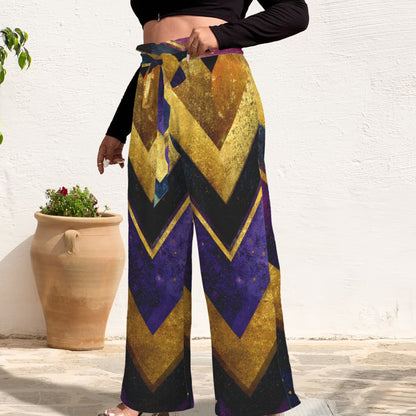 A person wearing high-waisted wide-leg pants with a bold chevron pattern in gold, purple, and black, paired with a black crop top and clear heels, standing against a white wall with potted plants.