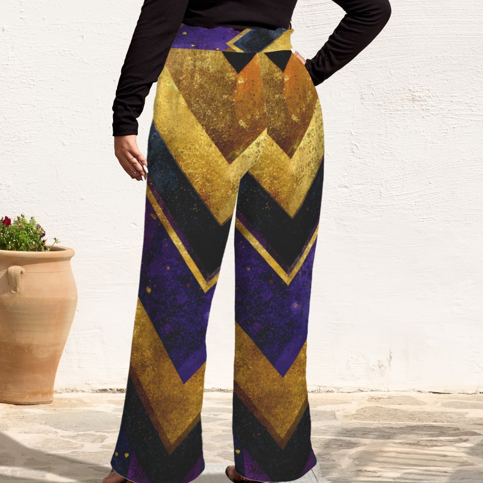 A person wearing wide-leg pants with a bold geometric pattern in purple, gold, and black, standing against a white wall next to a large terracotta pot.