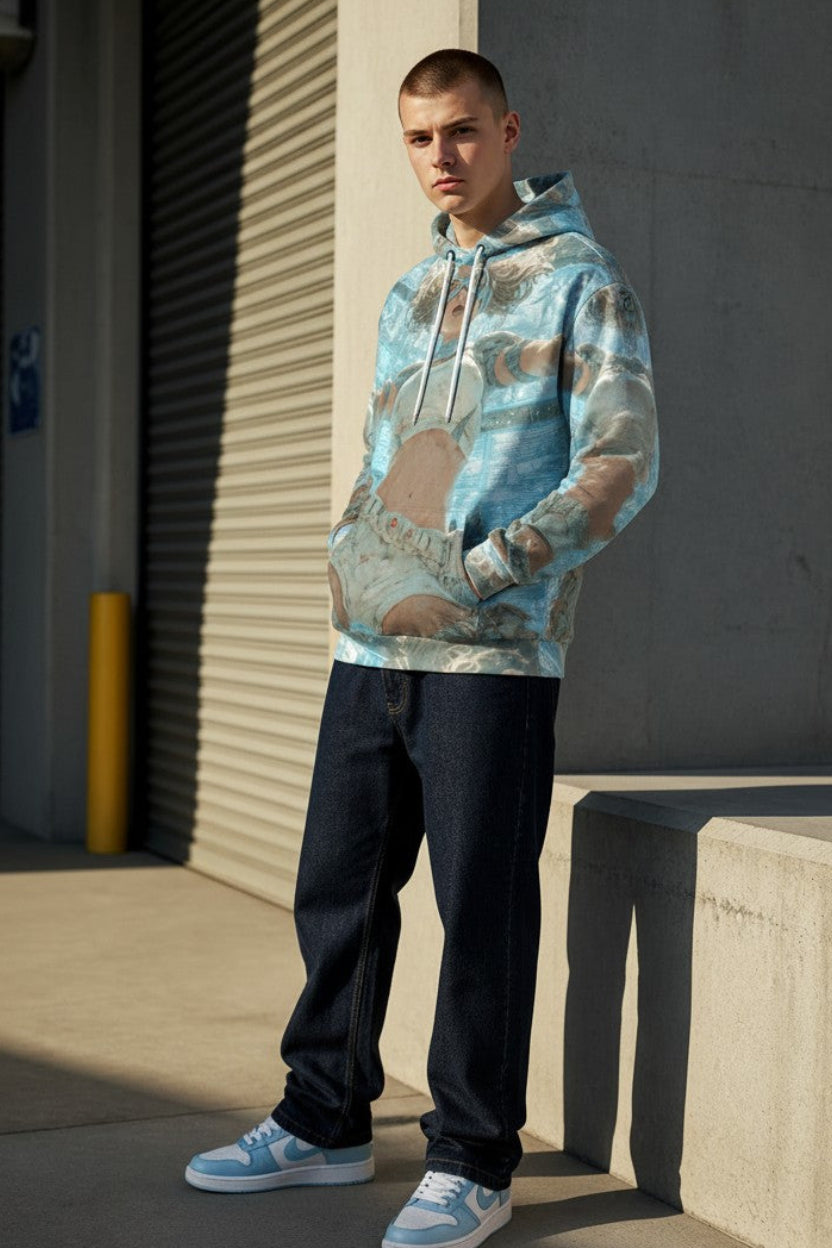 A man wearing a light blue and beige camouflage-patterned hoodie, dark jeans, and light blue sneakers stands outdoors against a concrete wall and metal roll-up door.