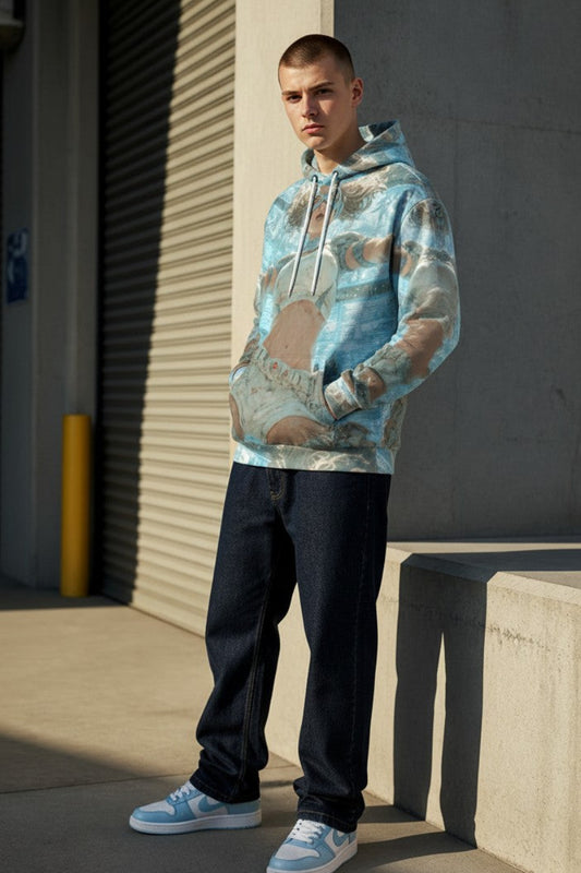 A man wearing a light blue and beige camouflage-patterned hoodie, dark jeans, and light blue sneakers stands outdoors against a concrete wall and metal roll-up door.