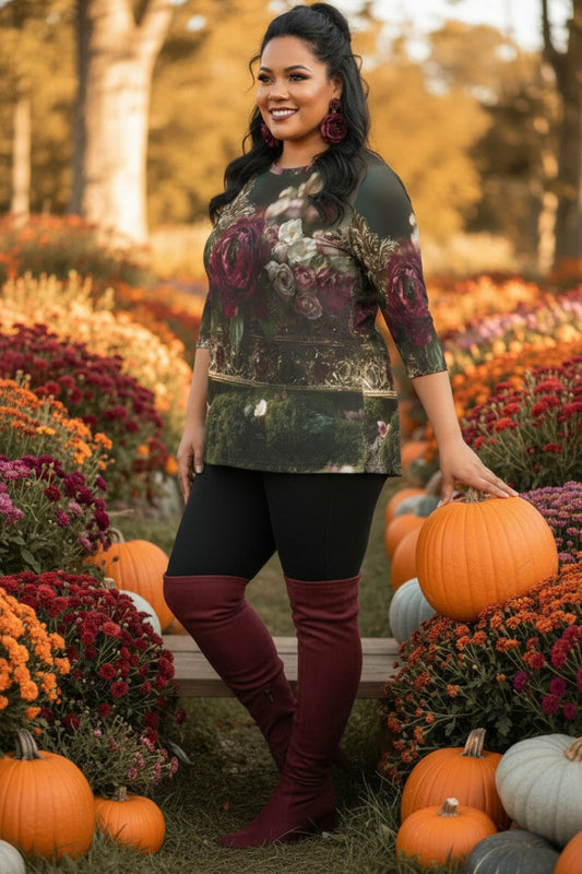 A woman stands in an autumn flower field surrounded by pumpkins, wearing a dark floral long-sleeve top, black leggings, and burgundy over-the-knee boots.