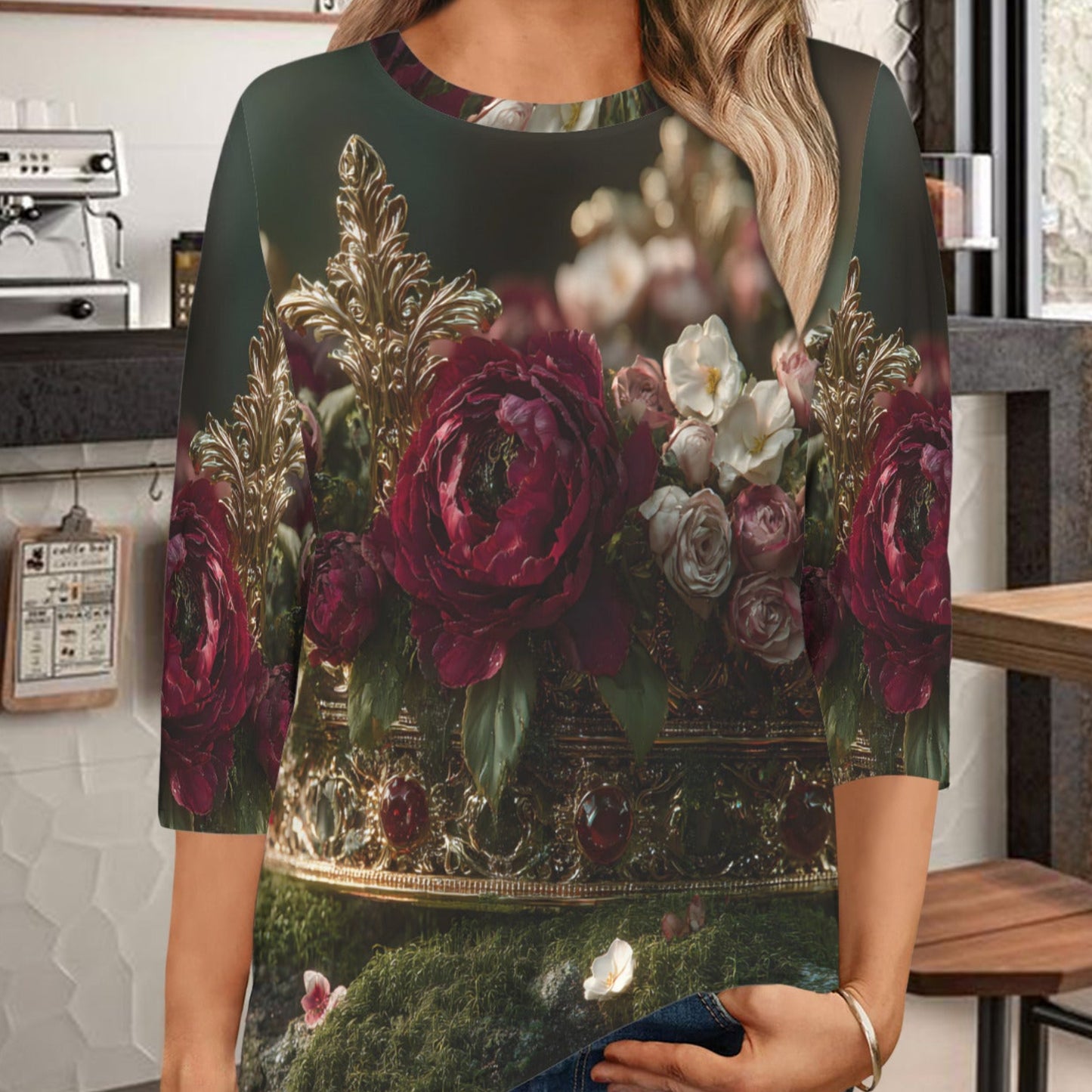 A woman wearing a three-quarter sleeve top with a dark green background and a large floral print featuring red and pink roses, photographed in a cafe setting.