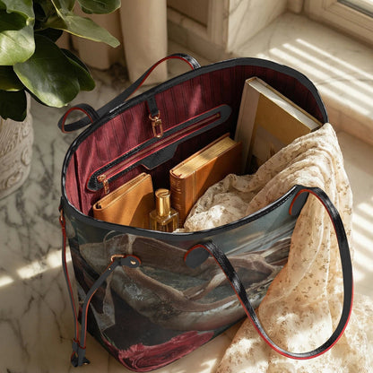 A tote bag with a dark blue and gray scenic print, red trim, and a burgundy interior, styled with books, a scarf, and a perfume bottle on a marble windowsill.