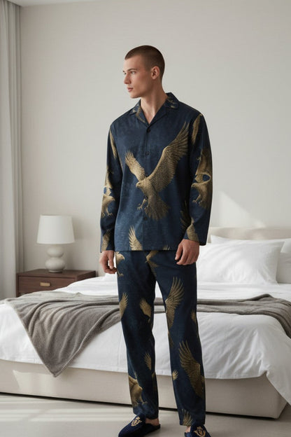 A man wearing a matching navy blue pajama set with a gold eagle print stands in a modern bedroom with a bed and neutral walls in the background.
