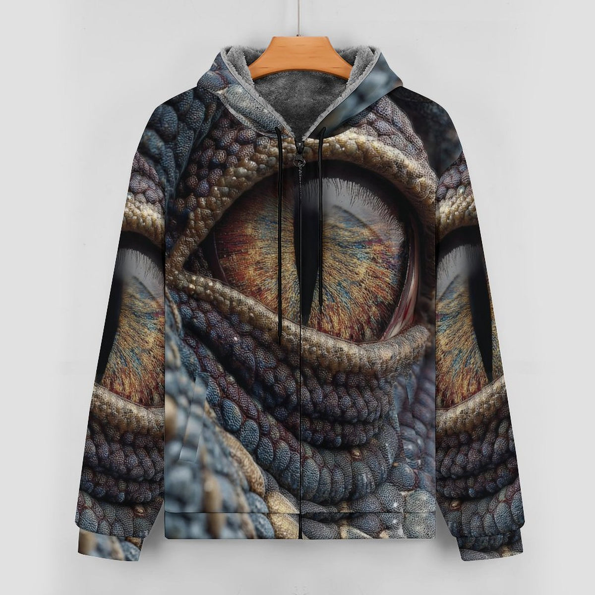 A hooded sweatshirt hanging on a wooden hanger against a light gray wall. The hoodie features an all-over printed design of a reptile or dragon eye with detailed scales and warm brown, gold, blue, and gray tones.