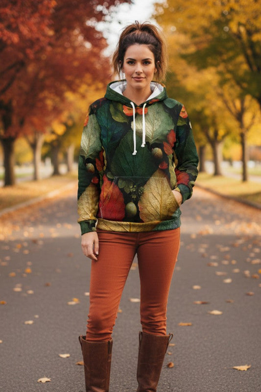 A woman stands on a paved path in an autumn park wearing a green and brown fall-themed hooded sweatshirt with a leaf print, rust-orange pants, and brown knee-high boots.