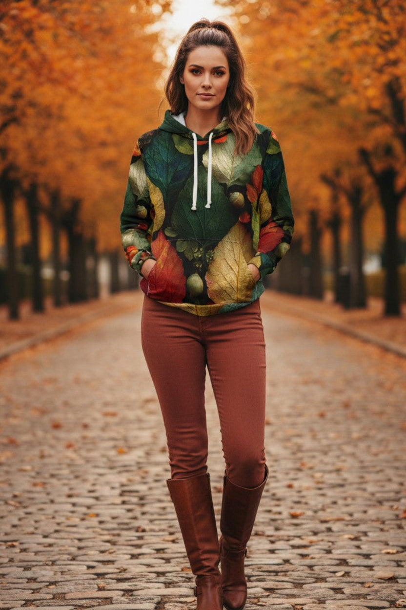 A woman stands on a cobblestone path lined with autumn trees, wearing a colorful leaf-print hoodie, rust-colored pants, and brown knee-high boots.