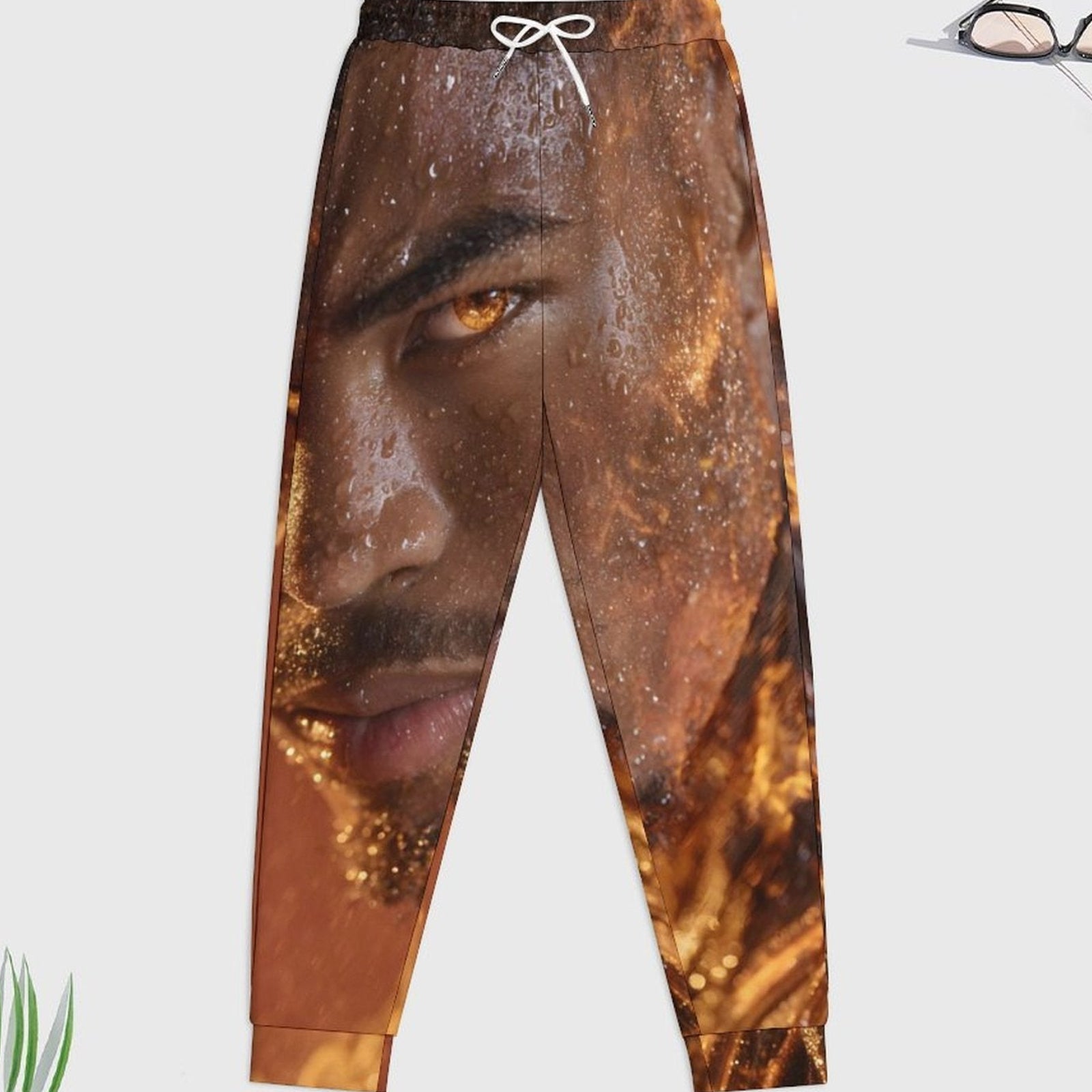 A pair of jogger-style pants with an all-over photographic print of a man's face in warm brown, gold, and orange tones, featuring a white drawstring waist and elastic cuffs, shown on a light gray background with small decorative plant and glasses props.