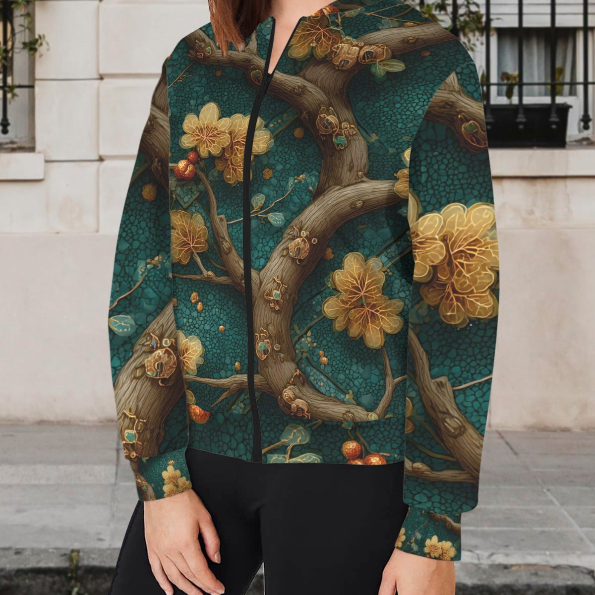 A woman wearing a hooded zip-up jacket with an intricate vintage-style floral and branch print in teal, gold, and brown, paired with black pants, standing outdoors in front of a light-colored stone building.