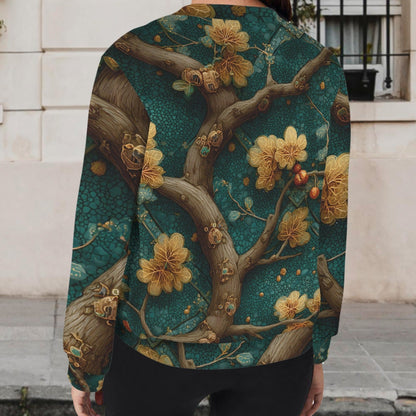 A woman viewed from behind wearing a long-sleeve bomber-style jacket with an intricate floral and branch print in teal, gold, and brown, standing outdoors in front of a light-colored building wall.