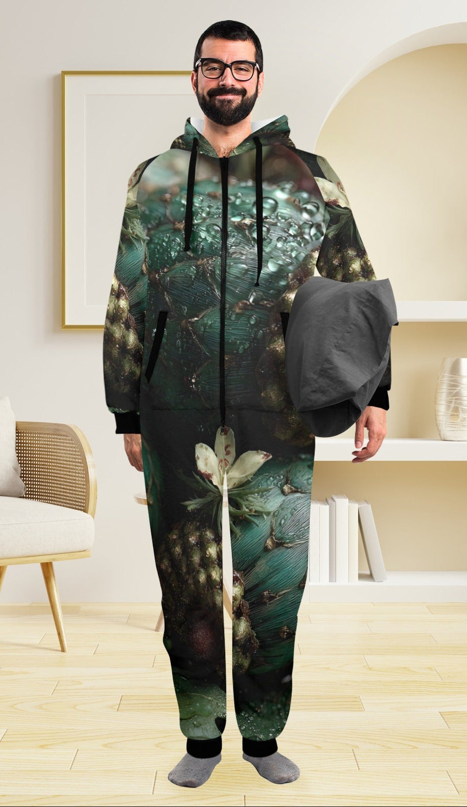 A man wearing a full-body hooded onesie with an abstract forest print in dark green, teal, black, and beige tones, standing in a bright modern room with a chair, pendant light, and shelving.