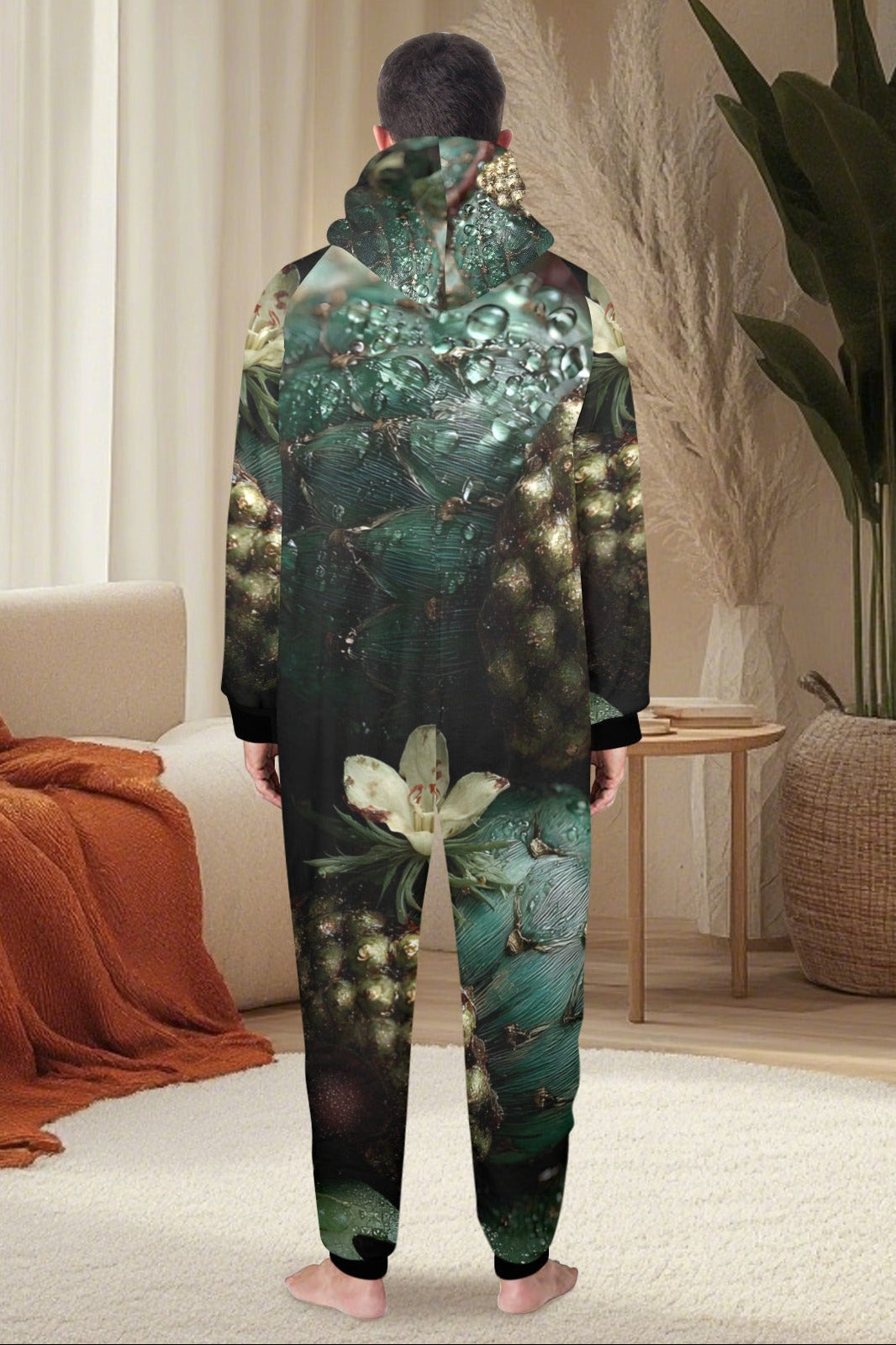 A person standing with their back to the camera wearing a full-body hooded onesie with an all-over nature print of green leaves, flowers, and glowing fireflies. The setting is a cozy, modern living room with a beige sofa, orange throw blanket, and a large potted plant.