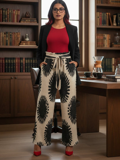 A woman stands in a library or study wearing a black blazer, a red top, and wide-leg cream pants with large black sunburst patterns. She is wearing red high heels, with bookshelves and a desk visible in the background.