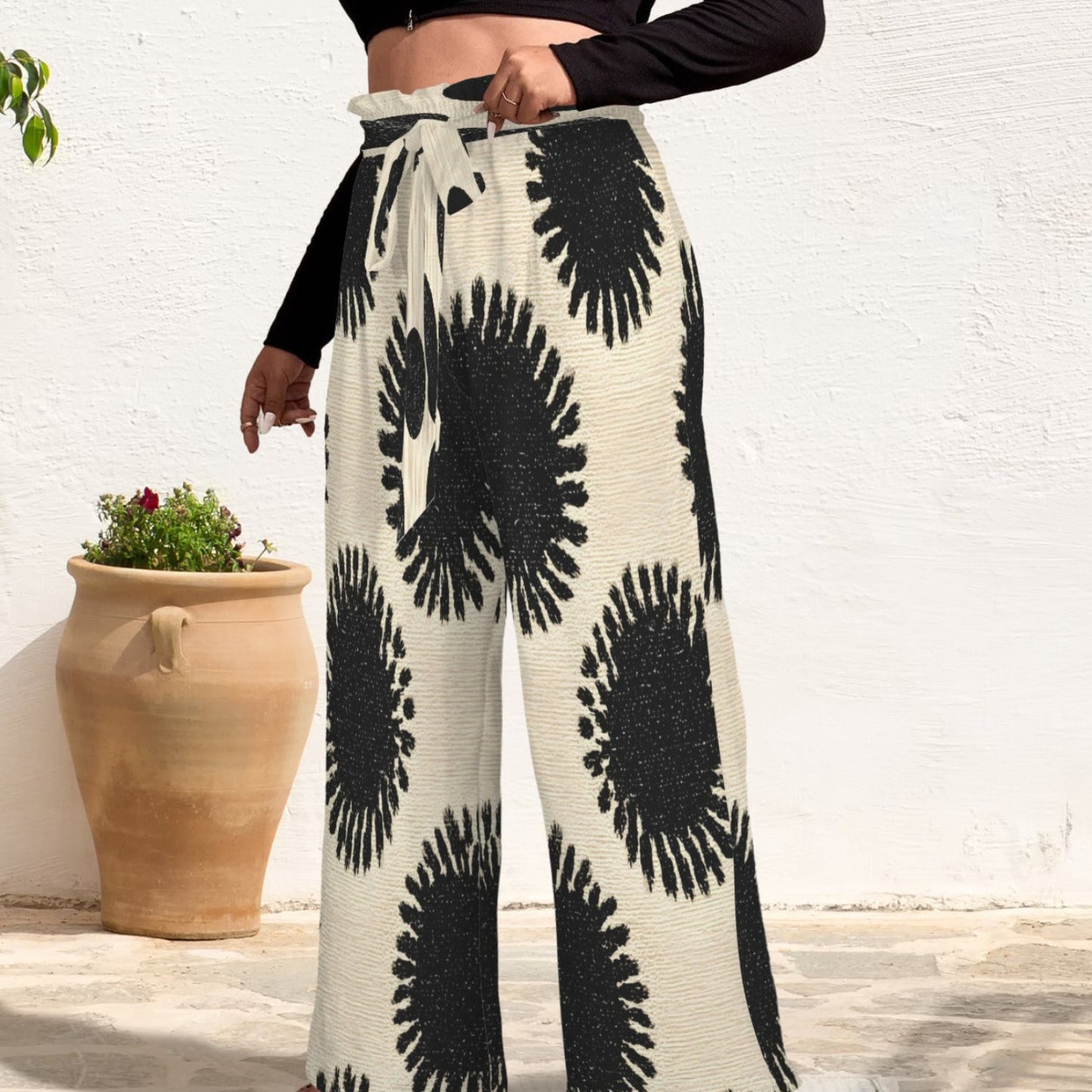 A person wearing high-waisted wide-leg pants with a black and off-white abstract floral pattern, paired with a black long-sleeve crop top and clear high-heel sandals, standing outdoors against a white wall.