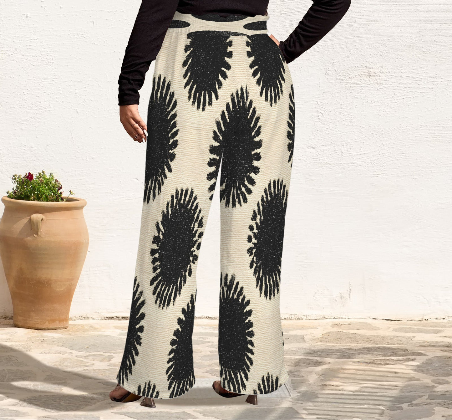 A person wearing high-waisted, wide-leg pants with a black and cream ikat-style circular pattern, paired with a black long-sleeve top, standing against a white stucco wall next to a large terracotta pot.