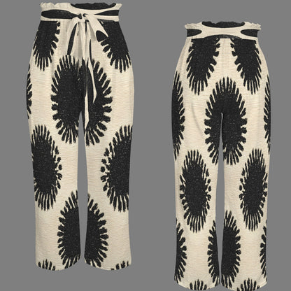 A pair of wide-leg knit pants shown from two angles on a solid gray background. The pants feature a cream-colored base with a bold black abstract circular pattern and a matching cream and black tie-waist belt.