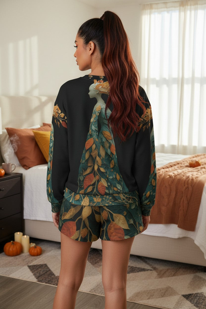 A woman stands in a bedroom wearing a matching black long-sleeve top and shorts set with a large autumn leaf print in teal, rust, and tan. She is shown from the back, with a bed, window, and pumpkins visible in the room.