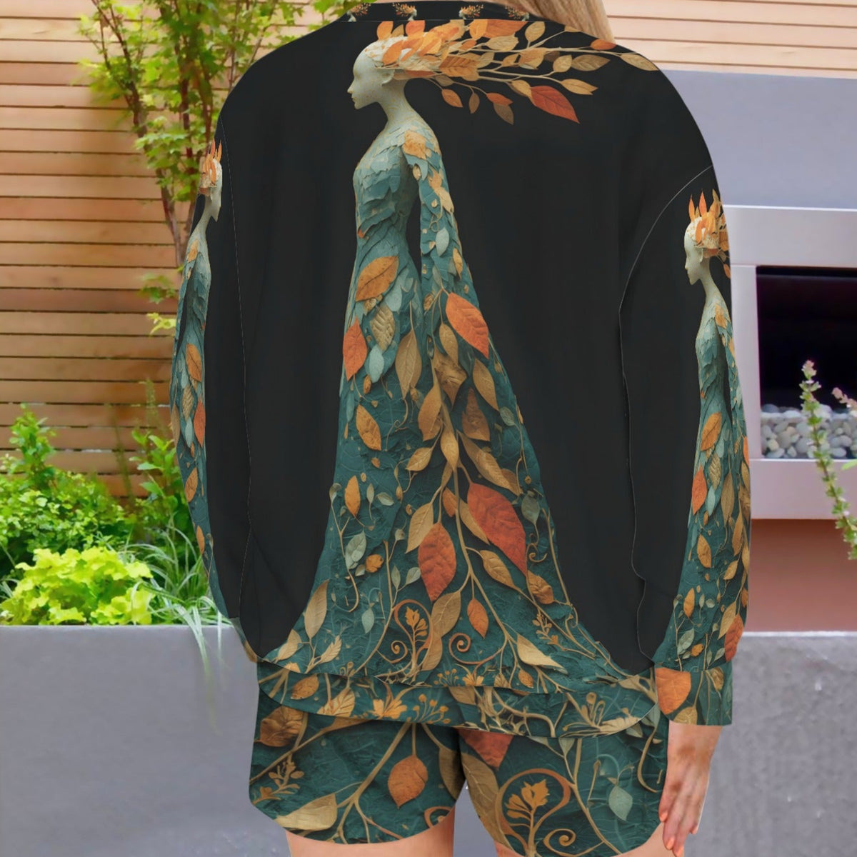 A woman viewed from behind wearing a black long-sleeve top and matching shorts with a large printed design of peacocks and autumn leaves in teal, gold, and rust tones. The setting is an outdoor patio with plants and a wooden wall.