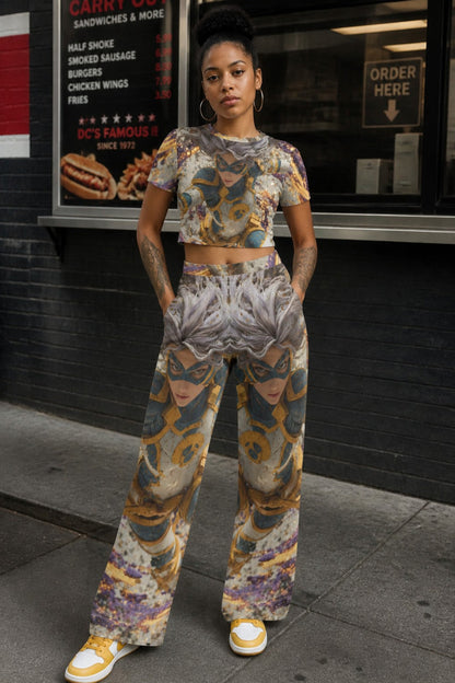 A woman stands on a city sidewalk in front of a restaurant window, wearing a matching cropped short-sleeve top and wide-leg pants set with an abstract multicolor print in gray, gold, blue, and purple tones. She is wearing yellow-and-white sneakers.