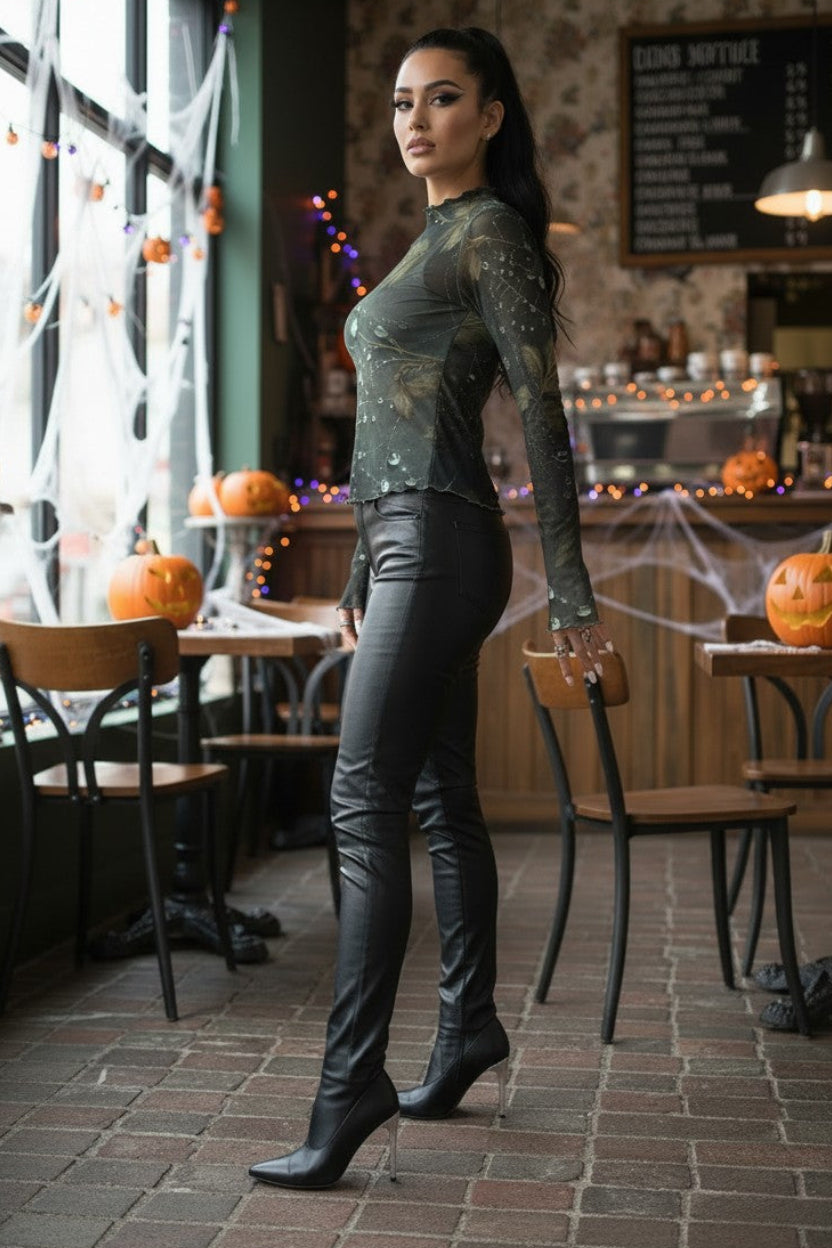 A woman stands in a Halloween-decorated cafe wearing a sheer long-sleeve black top with subtle glitter, black faux-leather pants, and black heeled boots.
