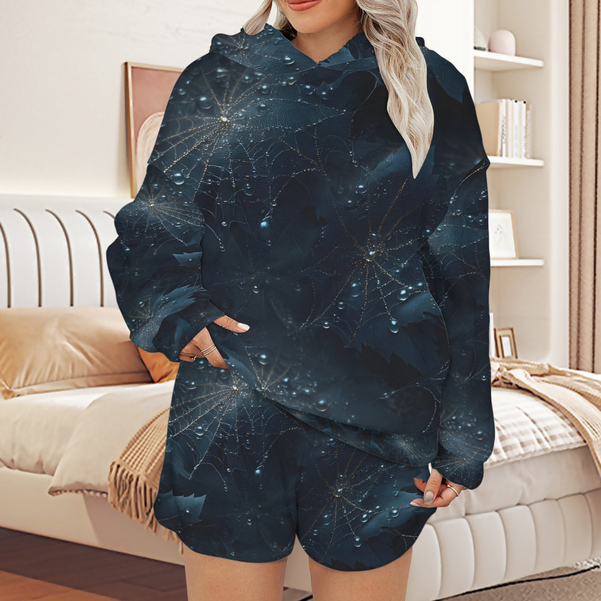 A woman wearing a matching dark blue hoodie and shorts set with an intricate celestial web pattern, standing in a modern bedroom setting.