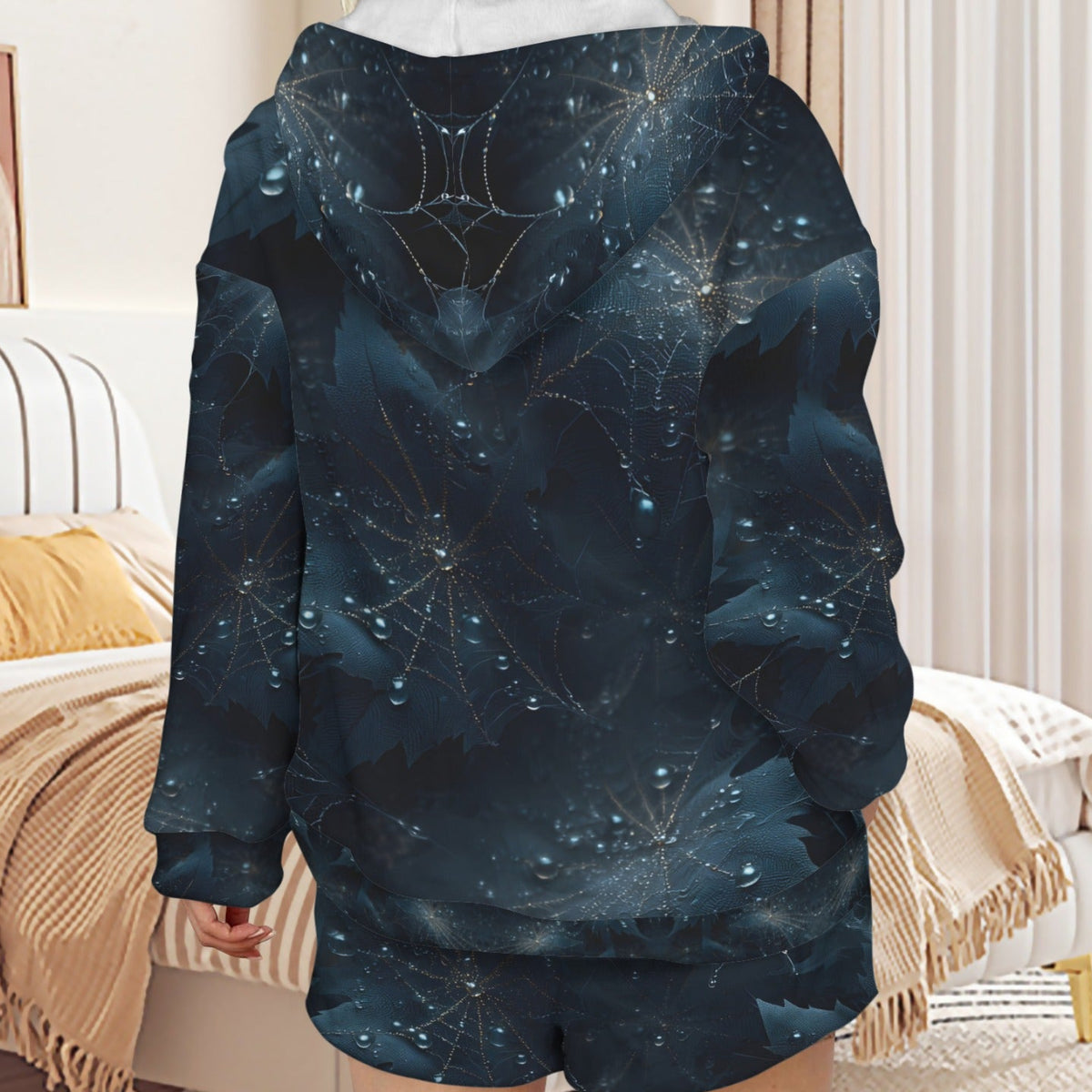 A person shown from the back wearing a dark hooded sweatshirt and matching shorts with an all-over celestial spiderweb print in deep navy, black, and blue tones with small glowing star-like highlights. The setting is a softly lit bedroom with a bed, pillows, curtains, and a rug visible.