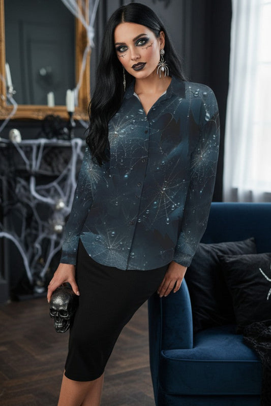 A woman models a long-sleeve button-up blouse with a dark blue and black spiderweb pattern, paired with black knee-length shorts, in a gothic-themed room with a blue velvet armchair and spider decorations.