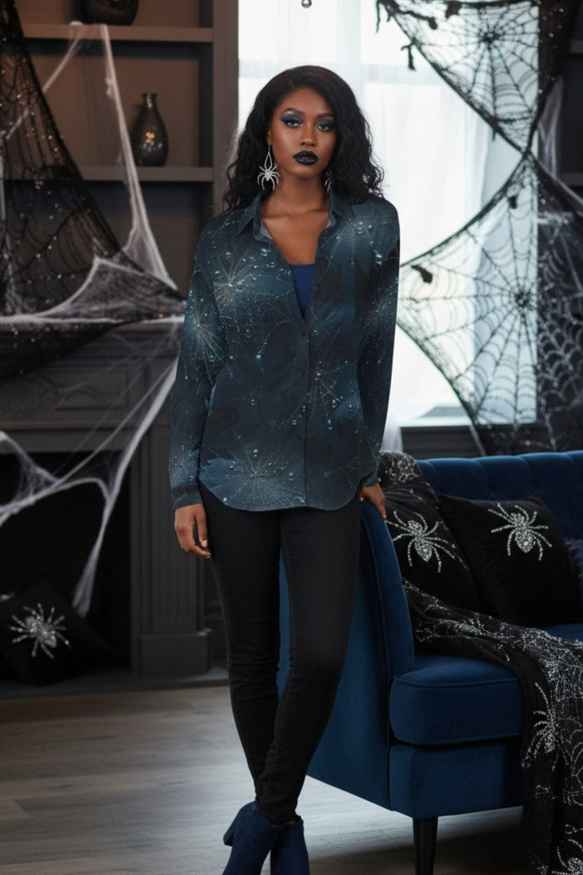 A woman stands in a Halloween-themed room wearing a long-sleeve button-up shirt with a dark blue and black galaxy print, black pants, and blue boots. The room features a blue velvet sofa with spider-themed pillows and blankets, and large spiderwebs hanging in the background.