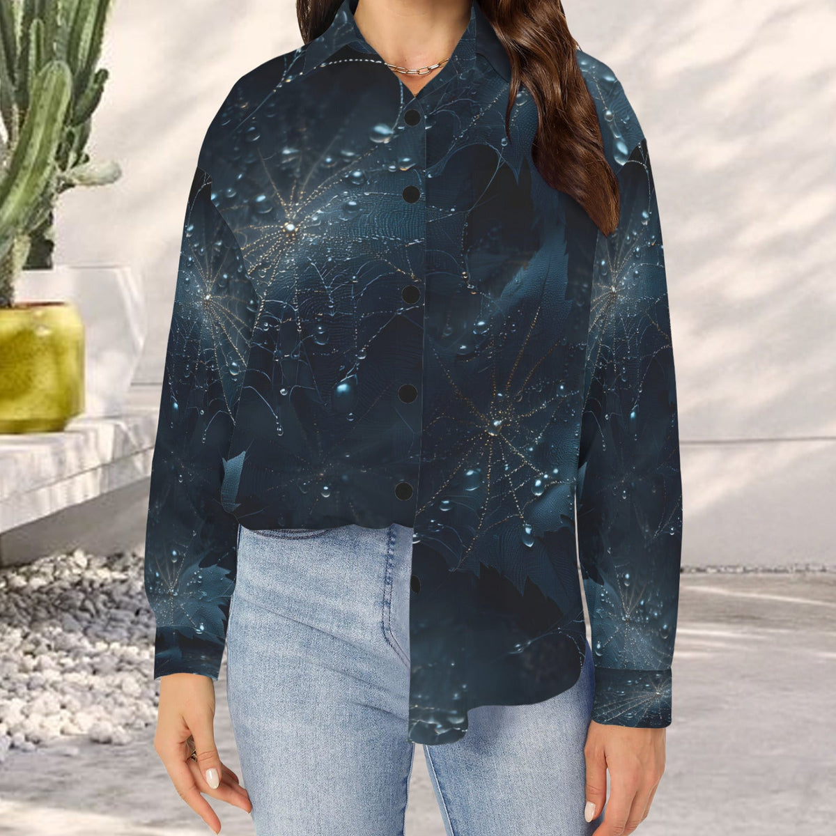 A woman wearing a long-sleeve button-up shirt with a dark blue and black spiderweb pattern, paired with light blue jeans, standing in a bright, modern outdoor setting.