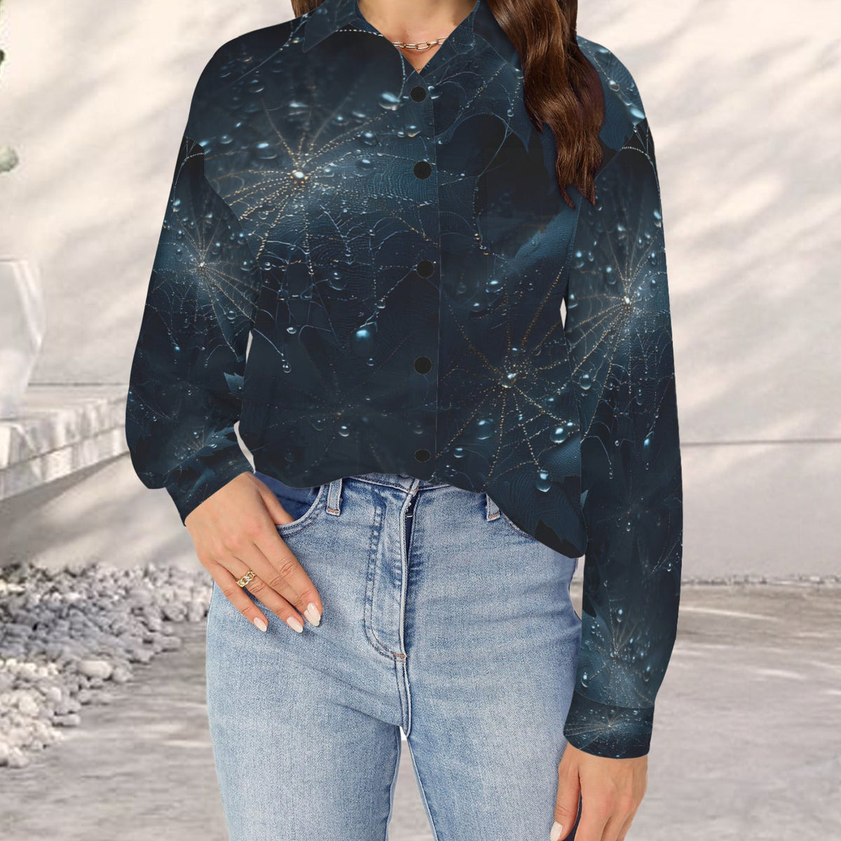 A woman wearing a long-sleeve button-up shirt with a dark blue and black celestial spiderweb pattern, paired with light blue jeans, standing in a modern outdoor setting.