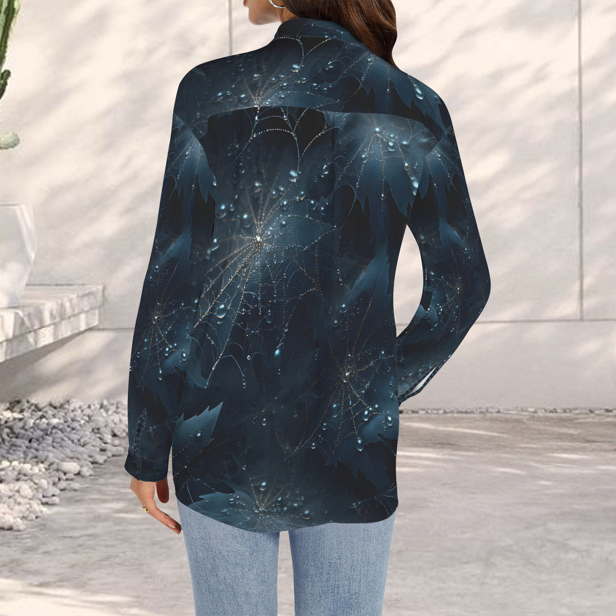 A woman shown from the back wearing a long-sleeve button-up shirt with a dark blue and black abstract fractal pattern accented by glowing light blue dots and web-like lines, paired with light blue jeans in an outdoor patio setting.
