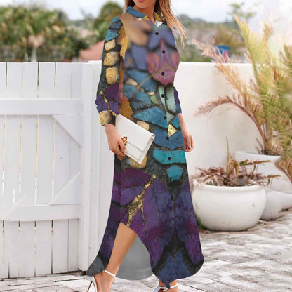 A woman wearing a long-sleeve, button-front maxi dress with an abstract multicolor print in purple, teal, blue, gold, and black, holding a white clutch and wearing white high-heeled sandals, standing outdoors on a paved walkway next to a white fence.