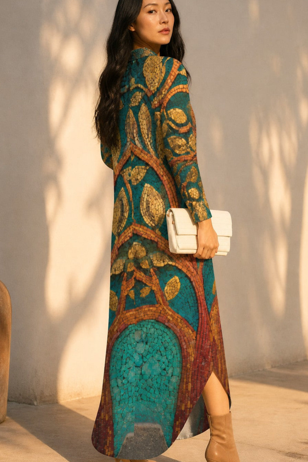 A woman wearing a long-sleeved maxi dress with a detailed tree pattern in teal, gold, and brown, holding a small white handbag and wearing beige ankle boots, standing against a sunlit beige wall with leaf shadows.