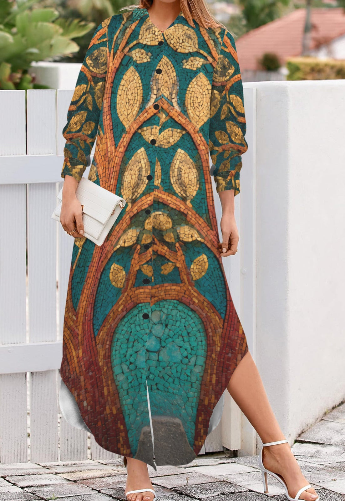 A woman wearing a long-sleeve maxi dress with a detailed botanical pattern in teal, gold, and brown, holding a white clutch and standing outdoors next to a white fence.